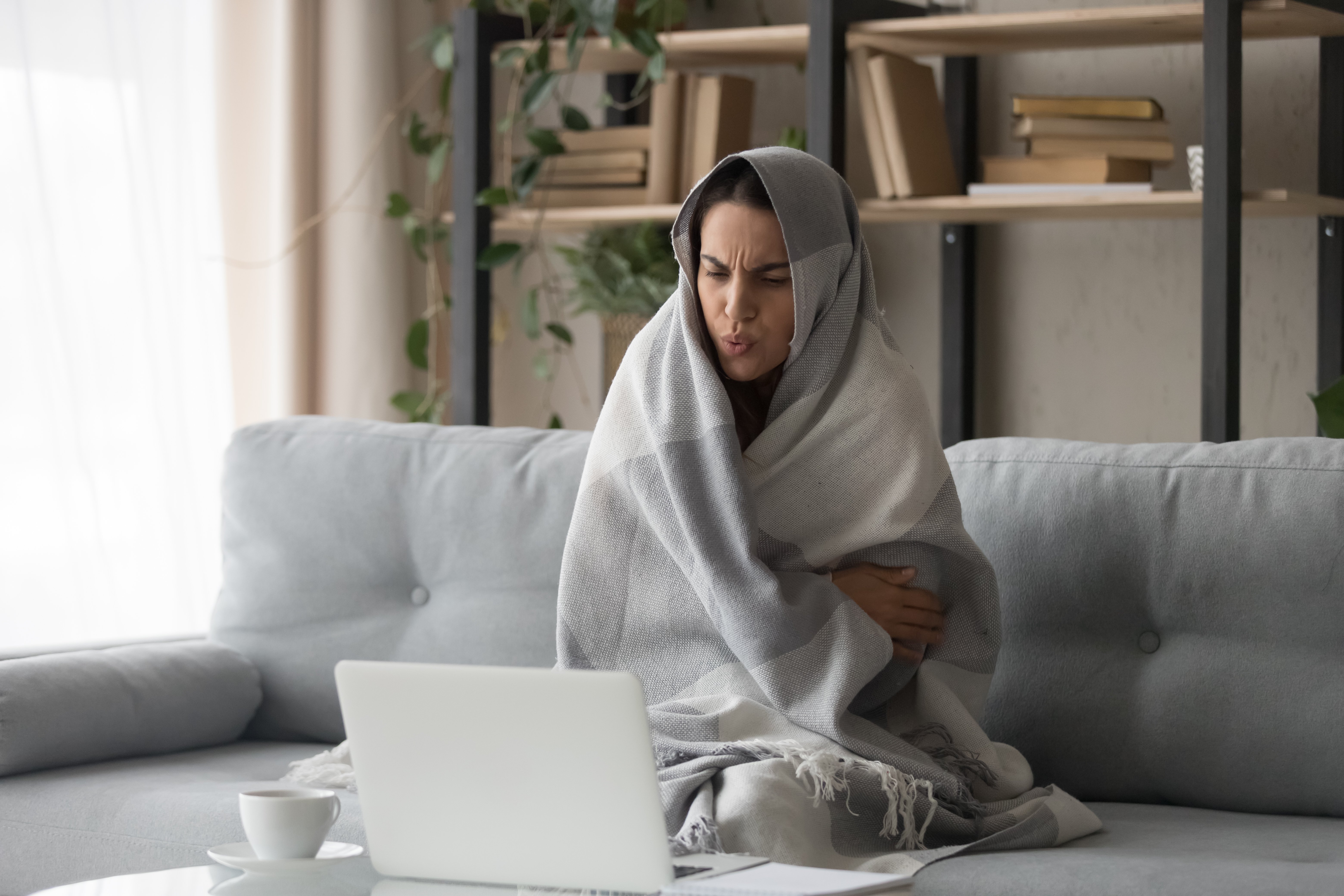 Woman on the couch covered by a blanket in front of her laptop with a cup of tea besides her.